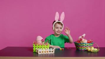 Young small kid placing bunny ears on his head in studio, preparing for easter sunday holiday celebration. Cheerful little boy sitting at a table to paint eggs and craft festive ornaments. Camera B. video