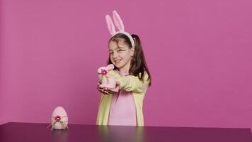 Smiling toddler with bunny ears showing her handmade easter ornaments, presenting a handcrafted decorated egg and rabbit. Young sweet schoolgirl holding festive decorations. Camera A. video