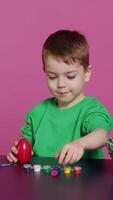 Vertical Video Young toddler focusing on decorating easter eggs with stamps and watercolor paint, preparing handcrafted ornaments for spring holiday festivity. Small cute boy coloring decorations. Camera A.