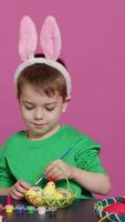 Vertical Video Happy small child arranging eggs and chick in a basket in preparation for easter sunday celebrations, creating festive decorations in studio. Little boy having fun coloring. Camera A.