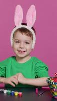 vertikal video ung små unge placering kanin öron på hans huvud i studio, framställning för påsk söndag Semester firande. glad liten pojke Sammanträde på en tabell till måla ägg och hantverk festlig ornament. kamera a.