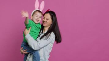 Mother lifting up her little boy and kissing him in studio, showing love and hugging cute kid against pink background. Sweet mom and son embracing each other and laughing. Camera A. video
