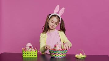 Excited young girl arranging painted eggs in a basket to prepare for easter holiday celebration, creating festive arrangements. Playful happy toddler with bunny ears, creative activity. Camera A. video