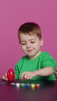 Vertical Video Ecstatic little kid using watercolor and paintbrushes to decorate eggs and create festive baskets as festive arrangements. Happy child enjoys painting and coloring decorations for easter. Camera A.