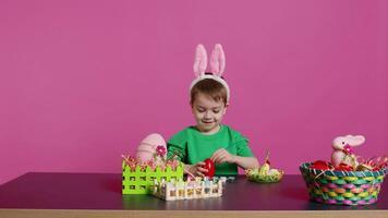 sorridente gioviale bambino in età prescolare pittura uova e ornamenti per Pasqua festa preparativi, utilizzando arte e mestiere materiale per decorare festivo disposizioni. giovane ragazzo avendo divertimento con cravatta tintura. telecamera b. video