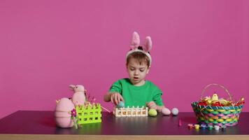 Happy toddler with bunny ears arranging basket filled with painted eggs, creating festive arrangements for the easter sunday celebration. Cheerful little kid enjoys art and craft. Camera B. video