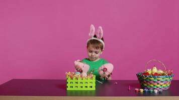 piccolo lieta bambino presentazione nascosto fatto a mano ornamenti creato nel preparazione di Pasqua vacanza celebrazione. giovane ragazzo con coniglietto orecchie mostrando dipinto decorazioni, un' coniglio e un uovo. telecamera b. video