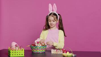 mignonne souriant bambin décorer une panier avec peint Pâques œufs, en présentant sa fabriqués à la main de fête arrangement dans studio. content charmant enfant création décorations et ornements. caméra un. video