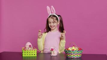 Small cheerful kid posing with fingers crossed in studio, sitting at a table filled with easter festive ornaments. Young schoolgirl hoping for luck and fortune during spring holiday celebration. Camera A. video