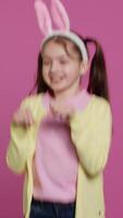 Vertical Video Joyful carefree schoolgirl jumping around in studio, imitating a rabbit and hopping against pink background. Cheerful active child wearing bunny ears and bouncing, adorable kid. Camera A.