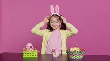 Smiling confident toddler placing bunny ears on her head and waving, saying hello against pink background. Cute joyful child creating decorations for easter sunday holiday event. Camera A. video