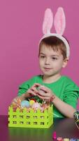 Vertical Video Excited little kid decorating baskets with eggs and grass to prepare for easter holiday festivity, creating festive arrangements. Cute toddler with bunny ears does craft activity. Camera A.