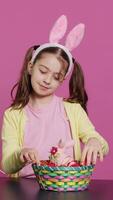 Vertical Video Excited young girl arranging painted eggs in a basket to prepare for easter holiday celebration, creating festive arrangements. Playful happy toddler with bunny ears, creative activity. Camera A.