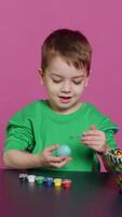 Vertical Video Little cheerful boy crafting handmade easter decorations by painting with watercolor and paintbrushes. Small young kid using art supplies coloring eggs for holiday, decorating activity. Camera A.