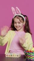 Vertical Video Joyful schoolgirl with bunny ears waving hello in front of camera, sitting at a table with festive decorations and arrangements for easter sunday celebration. Young excited child. Camera A.