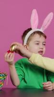 Vertical Video Sweet children knocking eggs together for easter tradition in studio, playing a seasonal holiday game against pink background. Lovely adorable kids having fun with festive decorations. Camera A.