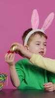 Vertical Video Sweet children knocking eggs together for easter tradition in studio, playing a seasonal holiday game against pink background. Lovely adorable kids having fun with festive decorations. Camera A.