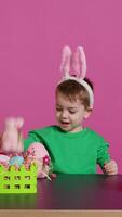 Joyful young kid playing around with festive painted decorations, showing a rabbit toy and a pink egg in front of camera. Smiling small boy with bunny ears having fun with ornaments. Camera B. video