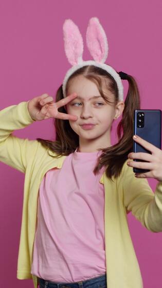 Vertical Video Confident happy young girl taking photos on smartphone webcam, feeling cute with her pigtails and bunny ears. Joyful smiling toddler takes pictures and fooling around in studio. Camera A. 42105136 Stock Video at Vecteezy-&gt;   