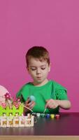 alegre pequeño niño pintura huevos para Pascua de Resurrección fiesta festividad en estudio, utilizando acuarela y Arte suministros. sonriente preescolar colorante festivo adornos en preparación para domingo. cámara b. video