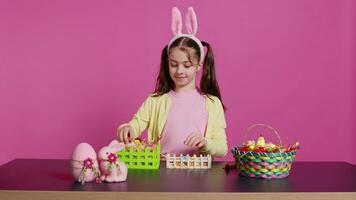 Smiling happy schoolgirl presenting a handmade decorated basket in studio, making easter holiday preparations against pink background. Young child showing handcrafted arrangements. Camera B. video