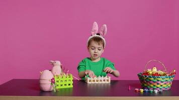 glücklich zufrieden Kind dekorieren Ostern Vereinbarungen mit handgemacht Artikel, arrangieren gemalt bunt Eier im ein Korb. froh wenig Junge feiern Frühling Fest, Rosa Hintergrund. Kamera b. video