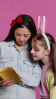 verticaal video moeder lezing sprookje verhaal naar haar zoet knap dochter, genieten van fictie boek met romans tegen roze achtergrond. vrolijk nieuwsgierig schoolmeisje met konijn oren luisteren naar haar mama. camera a.