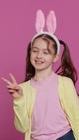 Vertical Video Young cheerful kid with pigtails showing peace sign in studio, feeling excited and confident about easter sunday celebration. Smiling youngster does signs against background. Camera A.