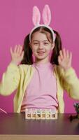 Energetic young girl with adorable bunny ears waving in studio, saying hello and greeting someone while she creates easter decorations. Joyful toddler posing against pink backdrop. Camera B. video