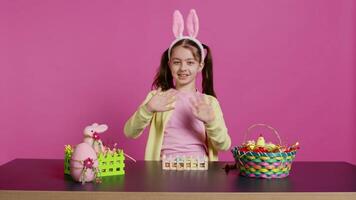 Joyful child putting bunny ears on her head and waving at camera, saying hello and smiling in excitement for the easter holiday. Happy toddler having fun with art and craft activity. Camera B. video