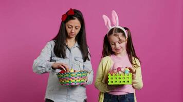 gioioso fiducioso bambino e madre mostrando Pasqua cesti su telecamera, decorazione festivo accordi per primavera vacanza. contento scolara con coniglietto orecchie in posa con sua mamma nel studio. telecamera b. video