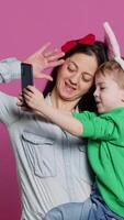 Vertical Video Lovely small child taking pictures with his mother on smartphone, trying to capture fun and cute moments against pink background. Little boy being playful and fooling around with phone. Camera A.