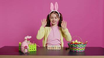 energisk ung flicka med förtjusande kanin öron vinka i studio, ordspråk Hej och hälsning någon medan hon skapar påsk dekorationer. glad litet barn Framställ mot rosa bakgrund. kamera b. video