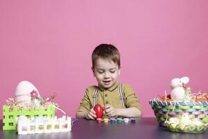 adorable pequeño joven haciendo encantador preparativos para Pascua de Resurrección domingo y pintura huevos con sellos y cepillos encantador niño gustos utilizando elaboración materiales a Decorar en estudio. foto