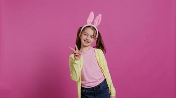 Young cheerful kid with pigtails showing peace sign in studio, feeling excited and confident about easter sunday celebration. Smiling youngster does signs against background. Camera B. video