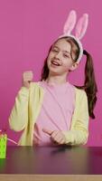 Happy schoolgirl with bunny ears showing thumbs up sign, feeling excited about easter holiday preparation. Cheerful child doing like okay sign and decorating baskets for festivity. Camera B. video