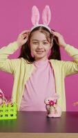 Smiling confident toddler placing bunny ears on her head and waving, saying hello against pink background. Cute joyful child creating decorations for easter sunday holiday event. Camera B. video