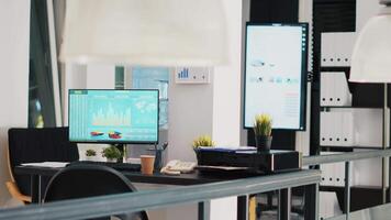 Office with computer monitor on desk showing stock exchange listings and display in background with business charts. PC in workspace with trading platform indexes and company data graphs in the back video