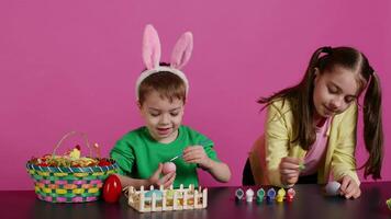 Young adorable kids coloring easter eggs with watercolor paint, creating lovely designs to celebrate spring holiday. Cheerful brother and sister painting festive decorations in studio. Camera B. video