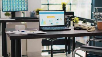 Wheelchair in office offering accessibility to workers with disability and laptop on desk showing business analytics with computer screen in background displaying stock exchange listings video