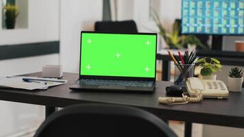 Office with green screen laptop on desk and monitor in background with stock exchange listings. Mockup notebook in workspace and computer screen in background displaying trading platform video