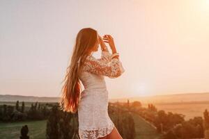 Happy woman in white boho dress on sunset in mountains. Romantic woman with long hair standing with her back on the sunset in nature in summer with open hands. Silhouette. Nature. Sunset. photo