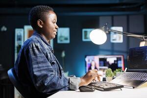 Female freelancer works at a desk, using a laptop to manage a futuristic information system. African american woman using her personal computer for cybersecurity and programming, working at home. photo