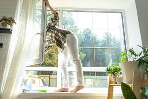 mujer a mano lavados el ventana de el casa con un trapo con rociar limpiador y fregona dentro el interior con blanco cortinas restaurar orden y limpieza en el primavera, limpieza servise foto