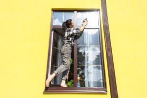 un mujer a mano lavados el ventana de el casa con un trapo con un rociar limpiador y un fregona afuera. la seguridad a altura, restaurar orden y limpieza en el primavera, limpieza Servicio foto