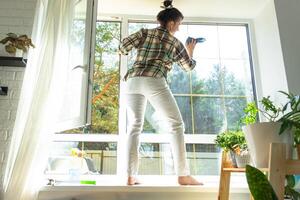 Woman manually washes the window of the house with a rag with spray cleaner and mop inside the interior with white curtains. Restoring order and cleanliness in the spring, cleaning servise photo