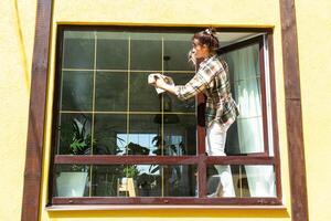 un mujer a mano lavados el ventana de el casa con un trapo con un rociar limpiador y un fregona afuera. la seguridad a altura, restaurar orden y limpieza en el primavera, limpieza Servicio foto