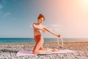 mujer mar pilates deportivo contento medio Envejecido mujer practicando aptitud en playa cerca mar, sonriente activo hembra formación con anillo en yoga estera afuera, disfrutando sano estilo de vida, armonía y meditación foto