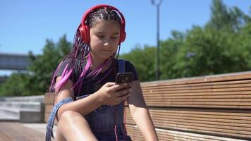 hermosa niña con largo coletas rosado color escuchando a música en auriculares utilizando teléfono inteligente sentado en un banco con un bueno estado animico en verano parque. lento movimiento. video