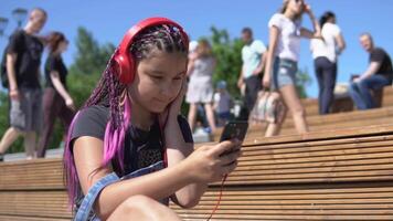 hermosa niña con largo coletas rosado color escuchando a música en auriculares utilizando teléfono inteligente sentado en un banco en verano parque. lento movimiento video